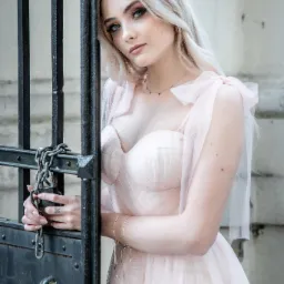 Portret kobiety w pudrowej sukni, opierającej się o metalową bramę z łańcuchem i kłódką; widoczny tatuaż motyla na dłoni.