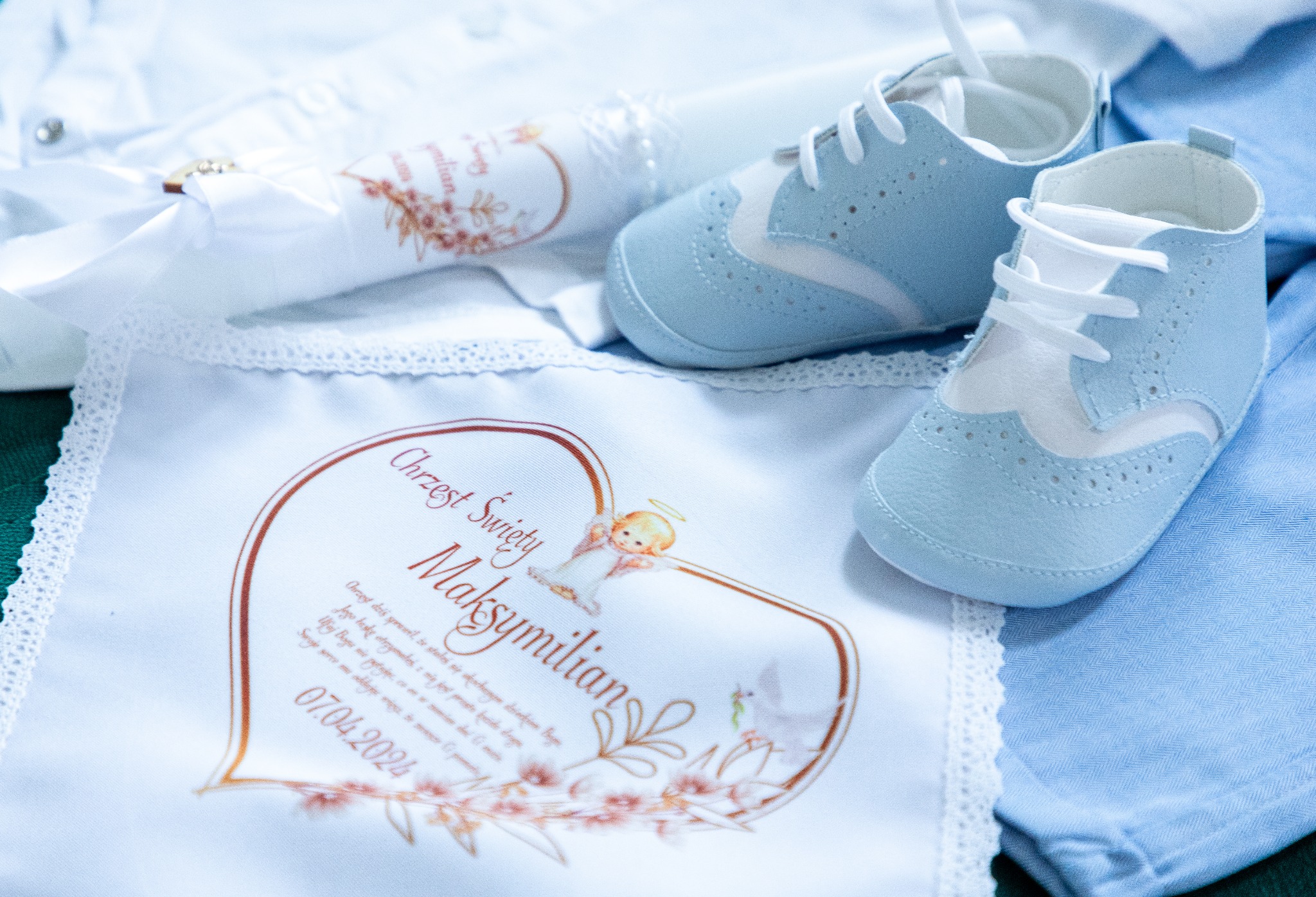 Kompozycja z błękitnych bucików niemowlęcych, świecy i serwetki z napisem 'Chrzest Święty Maksymilian 07.04.2024' oraz motywami kwiatowymi i aniołka, ułożona na jasnoniebieskiej tkaninie.