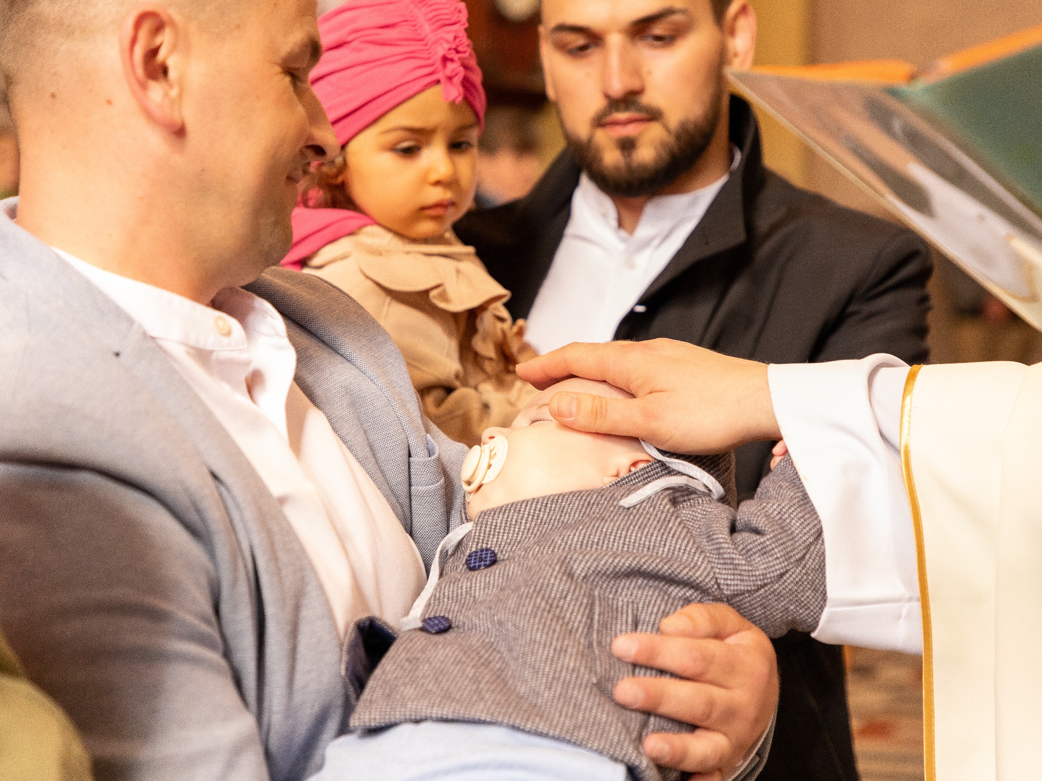 Ksiądz błogosławi śpiące niemowlę z smoczkiem na rękach ojca, w tle stoi dziewczynka w różowym turbanie i mężczyzna z brodą.