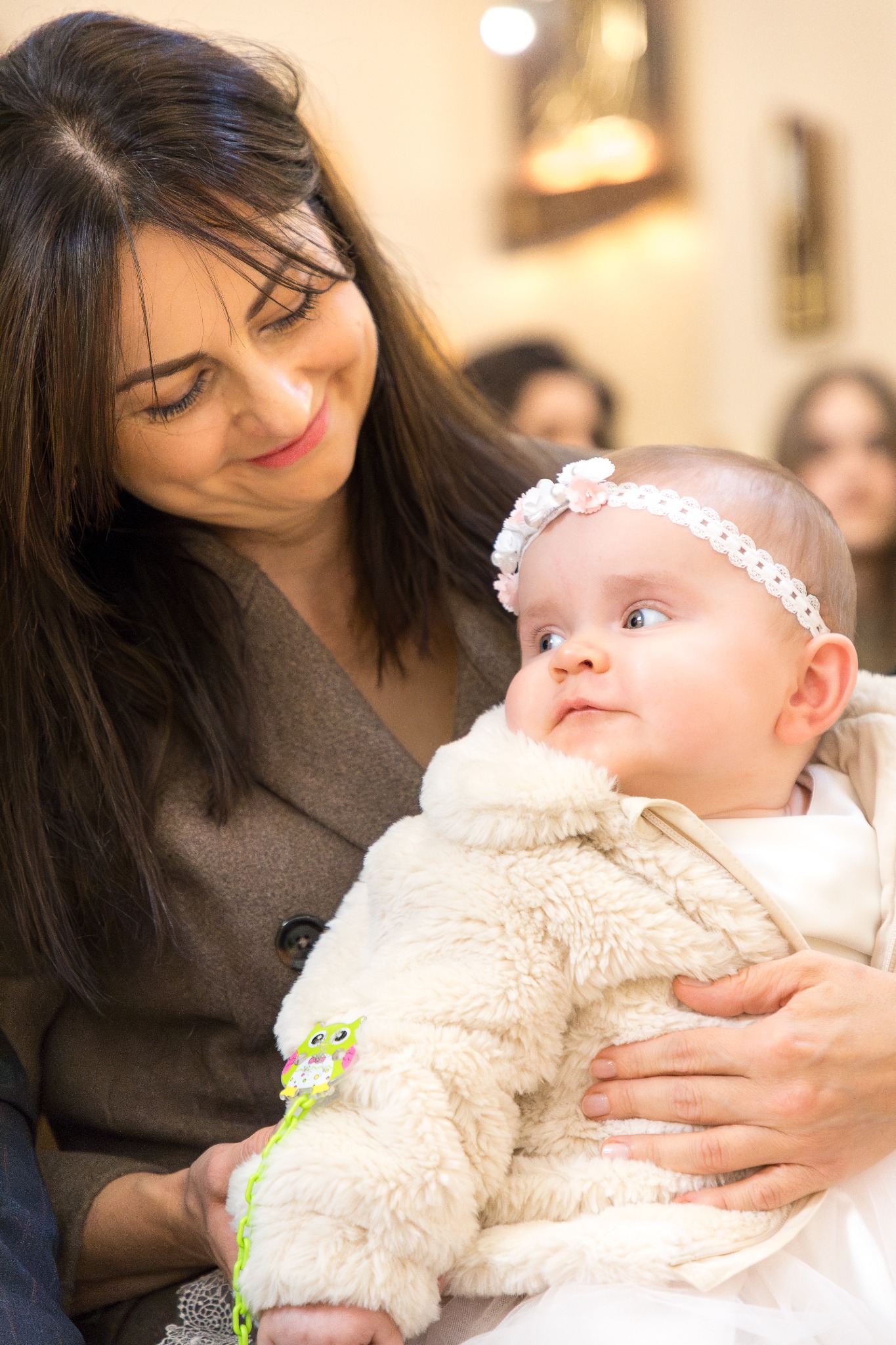 Mama trzymająca na rękach niemowlę ubrane w białe futerko z przypiętą sową, dziewczynka ma na głowie opaskę z kwiatami, w tle widoczne inne osoby.