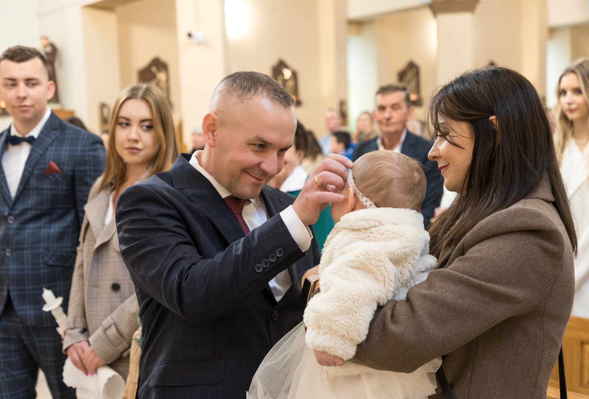 Ojciec poprawia opaskę niemowlęciu ubranemu w białe futerko i sukienkę podczas uroczystości w kościele, matka trzyma dziecko na rękach, w tle goście.