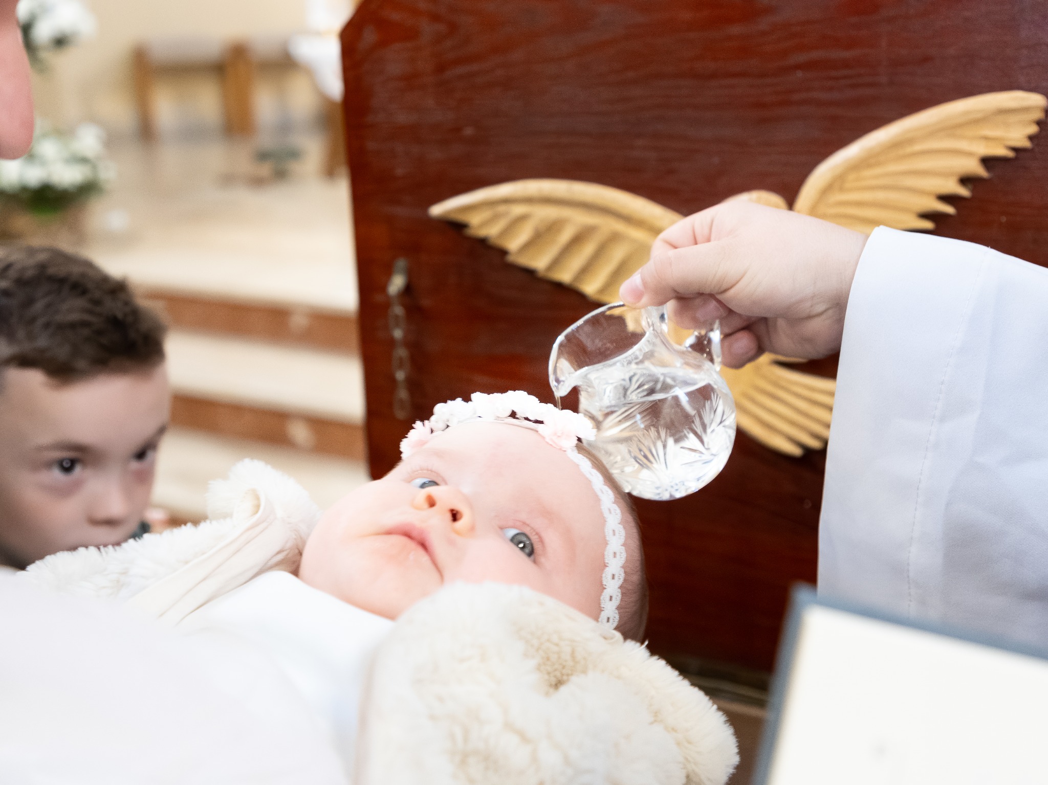 Chrzest dziecka w Lubartowie: Dziewczynka w białej czapce z kwiatami podczas ceremonii, w tle drewniany element dekoracyjny z wizerunkiem gołębicy.