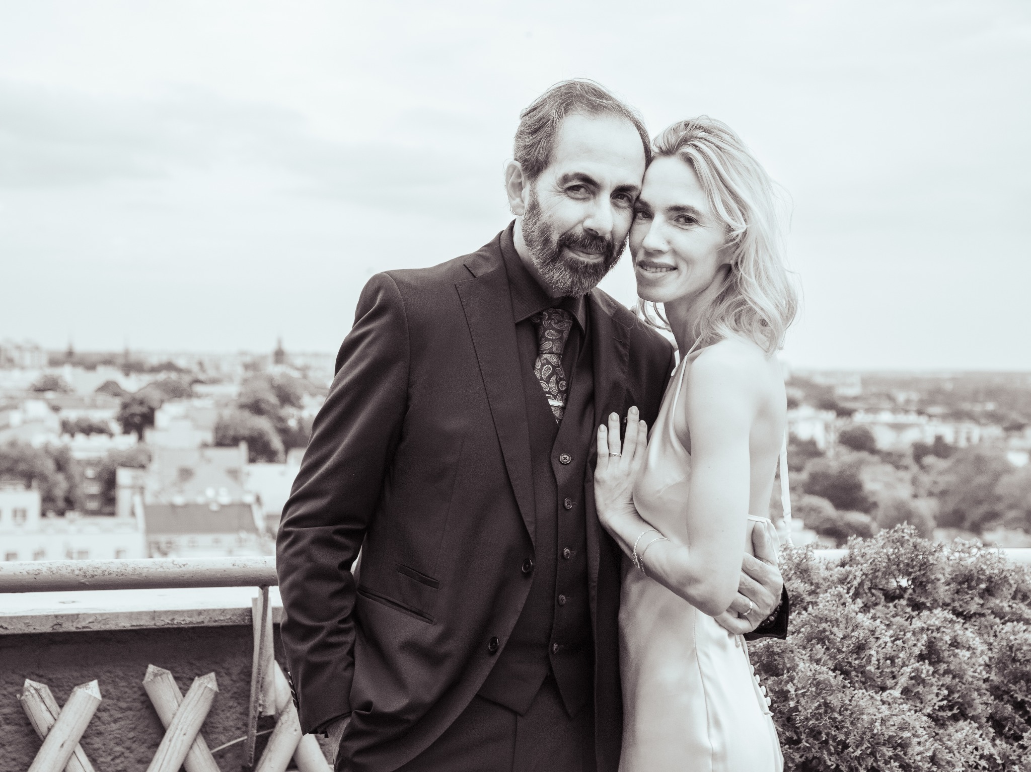 Czarno-białe zdjęcie pary: mężczyzna w garniturze i kobieta w sukience, obejmujący się na tle miejskiej panoramy z zielenią. Kobieta opiera dłoń na ramieniu mężczyzny.