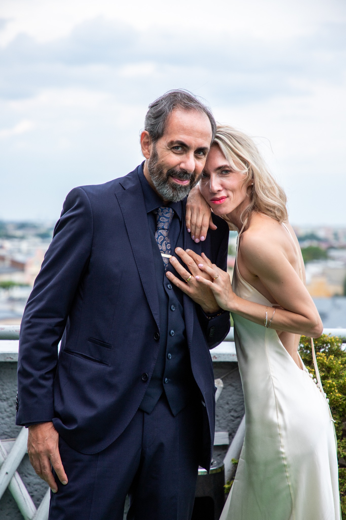 Elegancka para na tle miejskiej panoramy: mężczyzna w granatowym garniturze i kobieta w białej sukni, pozujący na tle nieba i zabudowań. Uśmiechy, bliskość, naturalne światło.