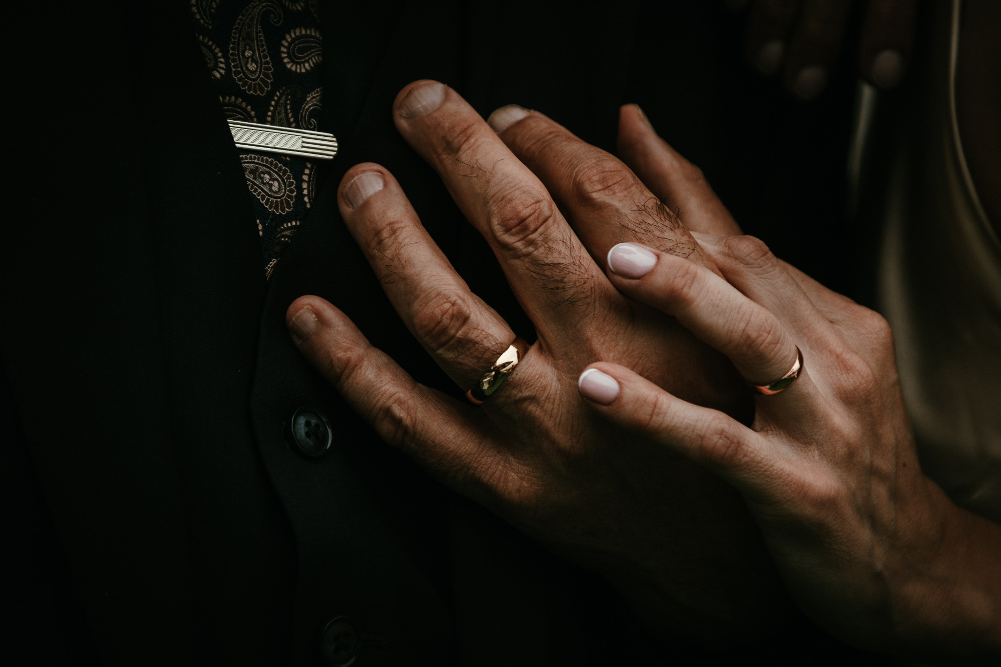 Dłonie pary młodej z złotymi obrączkami na tle ciemnej kamizelki z wzorem paisley i spinki do krawata. Delikatne światło podkreśla detale.