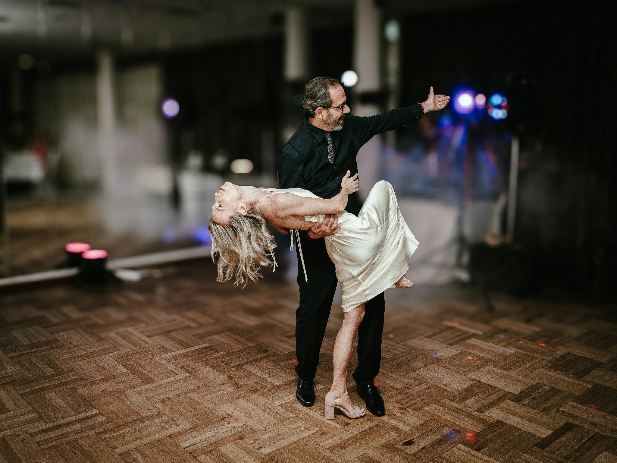 Para tańczy na parkiecie podczas uroczystości. Mężczyzna w czarnym stroju trzyma kobietę w białej sukni w pozycji tanecznej, w tle widoczne światła i dekoracje.