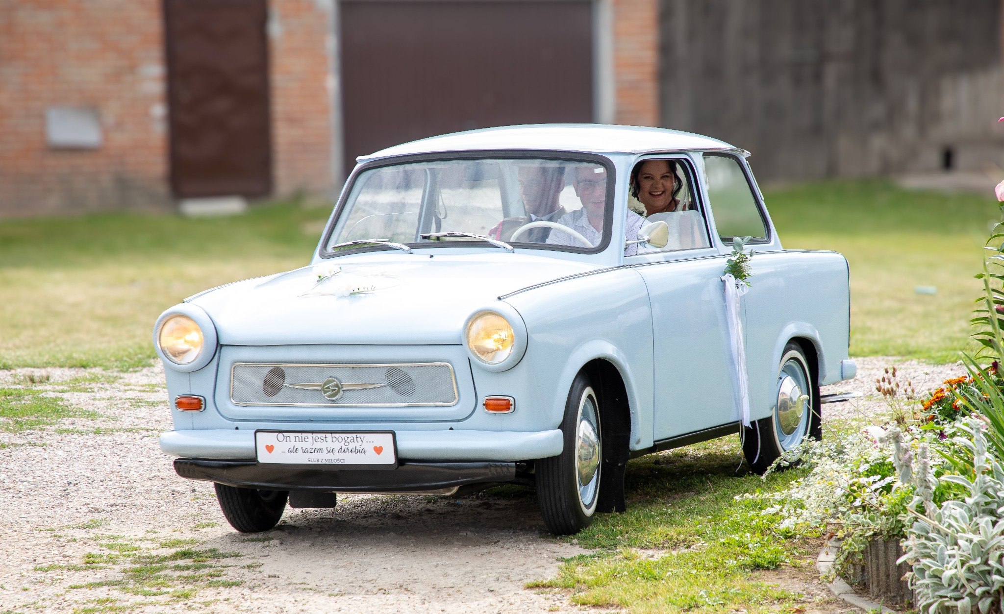 Błękitny Trabant z dekoracjami ślubnymi, z uśmiechniętą parą młodą w środku, na tle zieleni i ceglanego budynku. Tablica rejestracyjna z napisem 'Ślub z miłości'.