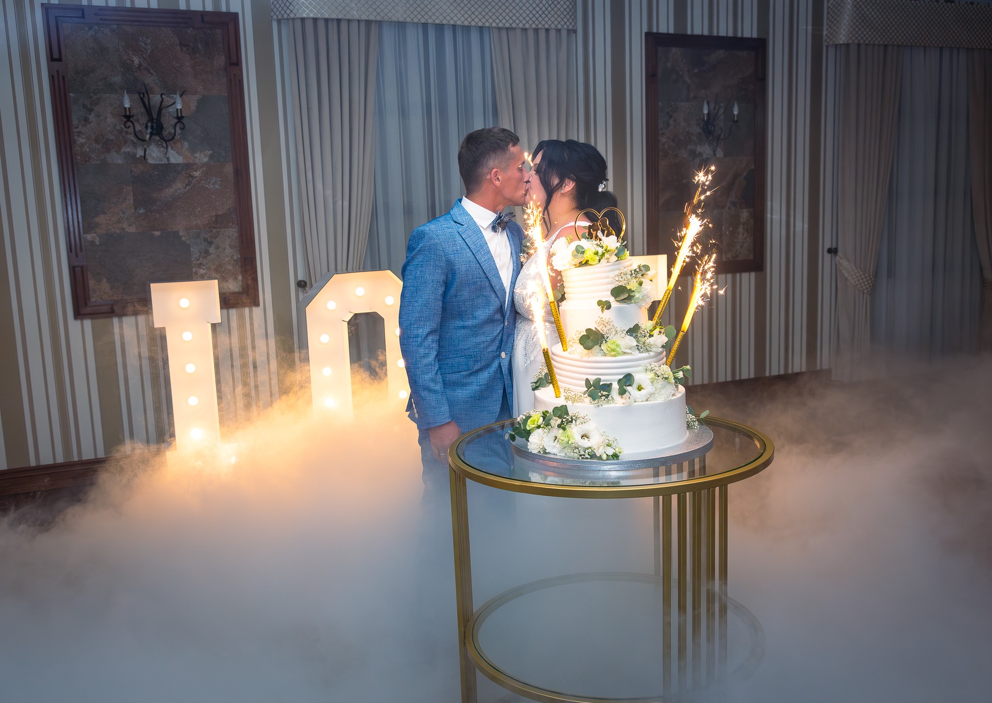 Pocałunek pary młodej za tortem weselnym z zimnymi ogniami, na tle dekoracji świetlnych i efektu dymu. Pan młody w niebieskiej marynarce.