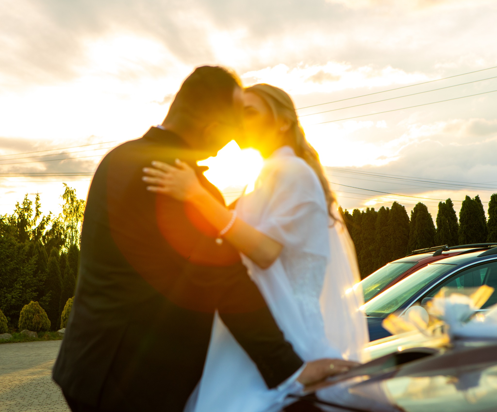 Para młoda w objęciach na tle zachodzącego słońca, zarys aut w tle. Romantyczna atmosfera, ciepłe światło.
