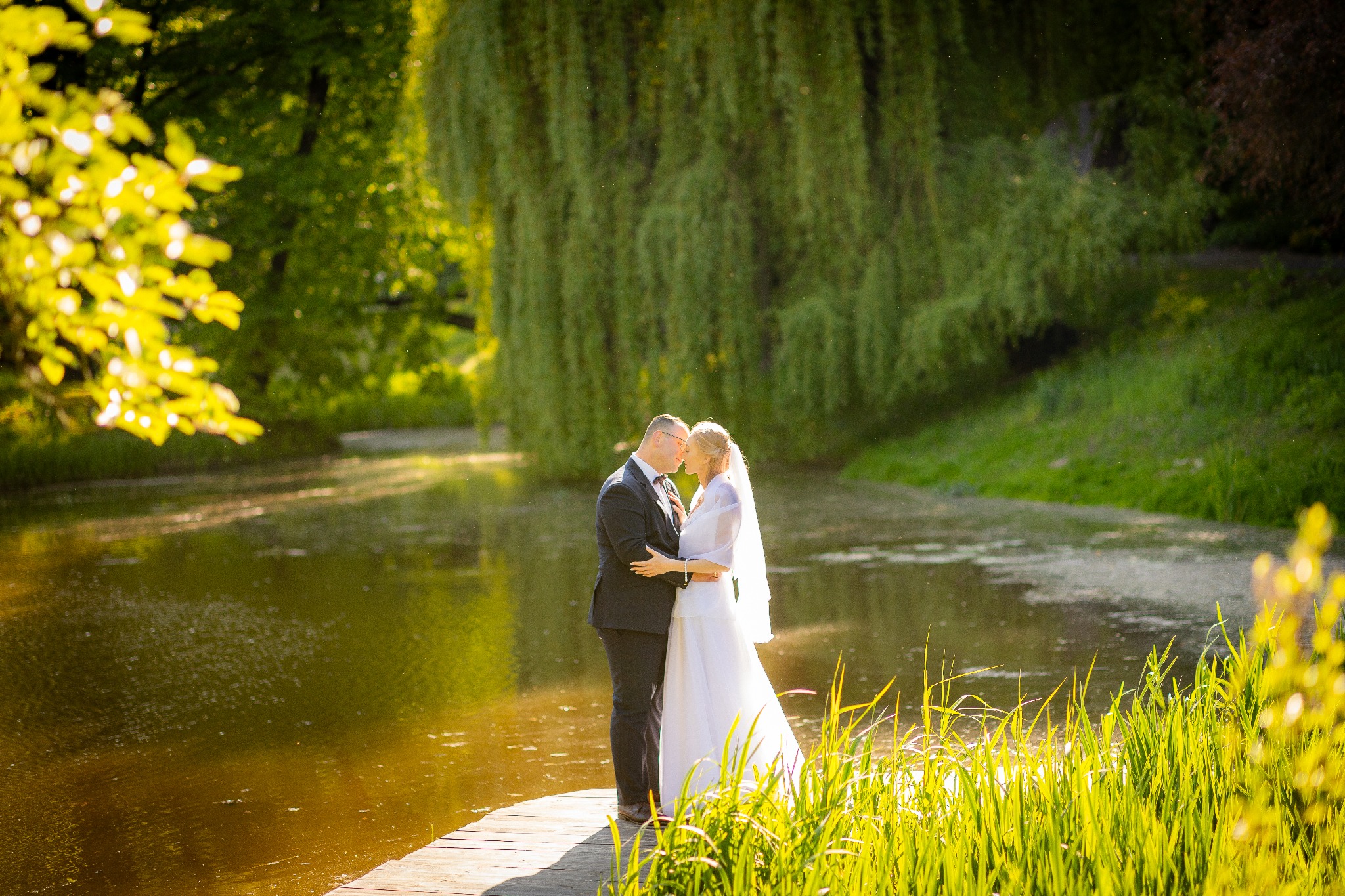 Para młoda w objęciach na drewnianym pomoście nad stawem, w tle malowniczy, zielony krajobraz z drzewami i odbiciami w wodzie, w ciepłym, naturalnym oświetleniu.