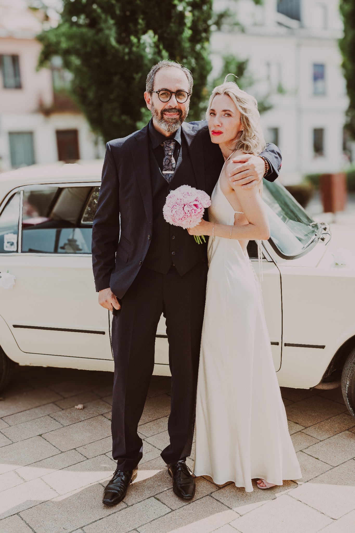 Para młoda w eleganckich strojach pozuje przy kremowym, zabytkowym samochodzie z kwiatami. Pan młody w okularach, panna młoda z bukietem różowych piwonii.