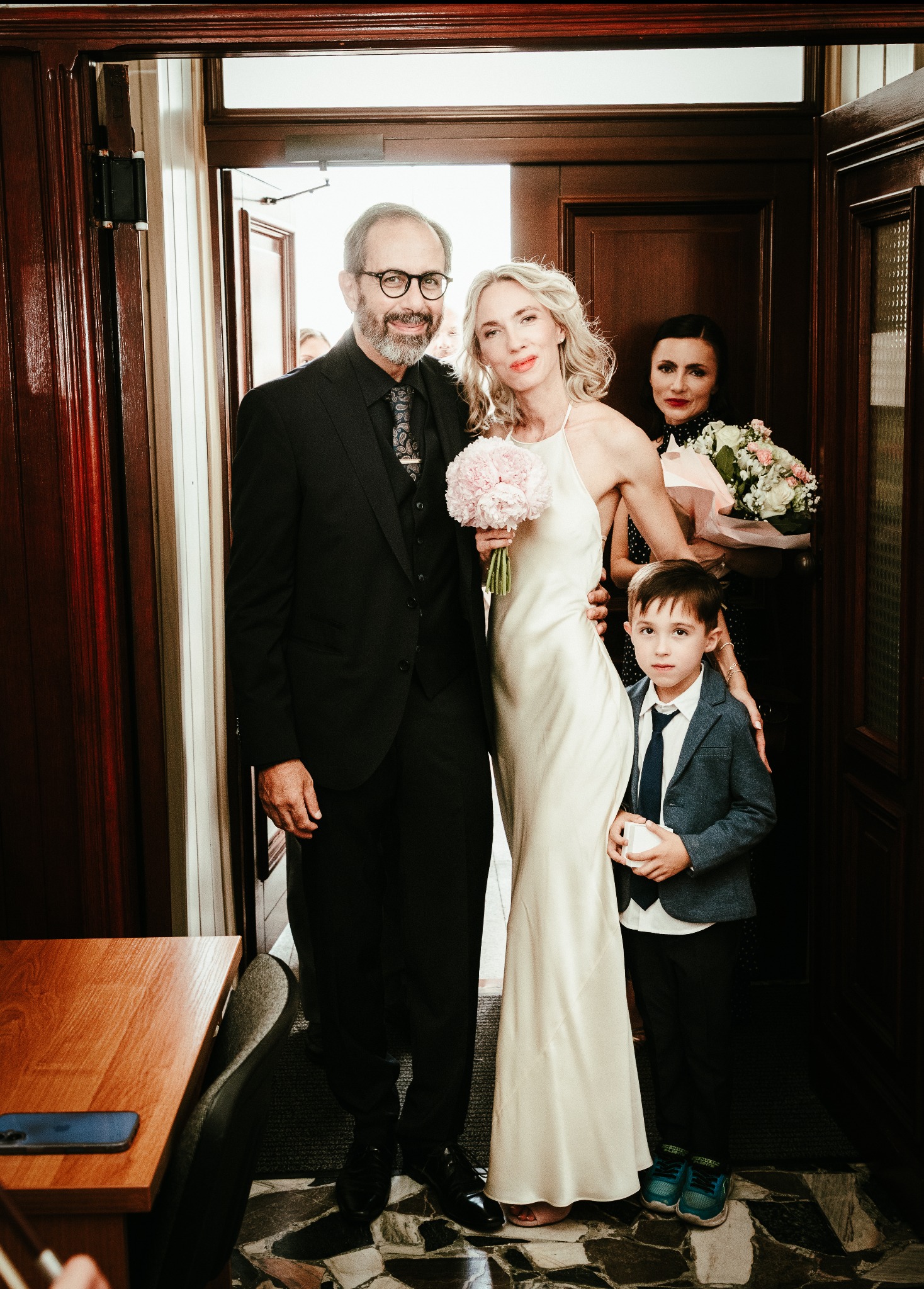 Elegancka para młoda z dzieckiem i kobietą z bukietem, pozująca w drzwiach. Panna młoda w satynowej sukni, pan młody w czarnym garniturze. Uśmiechnięte twarze, ciepłe oświetlenie.