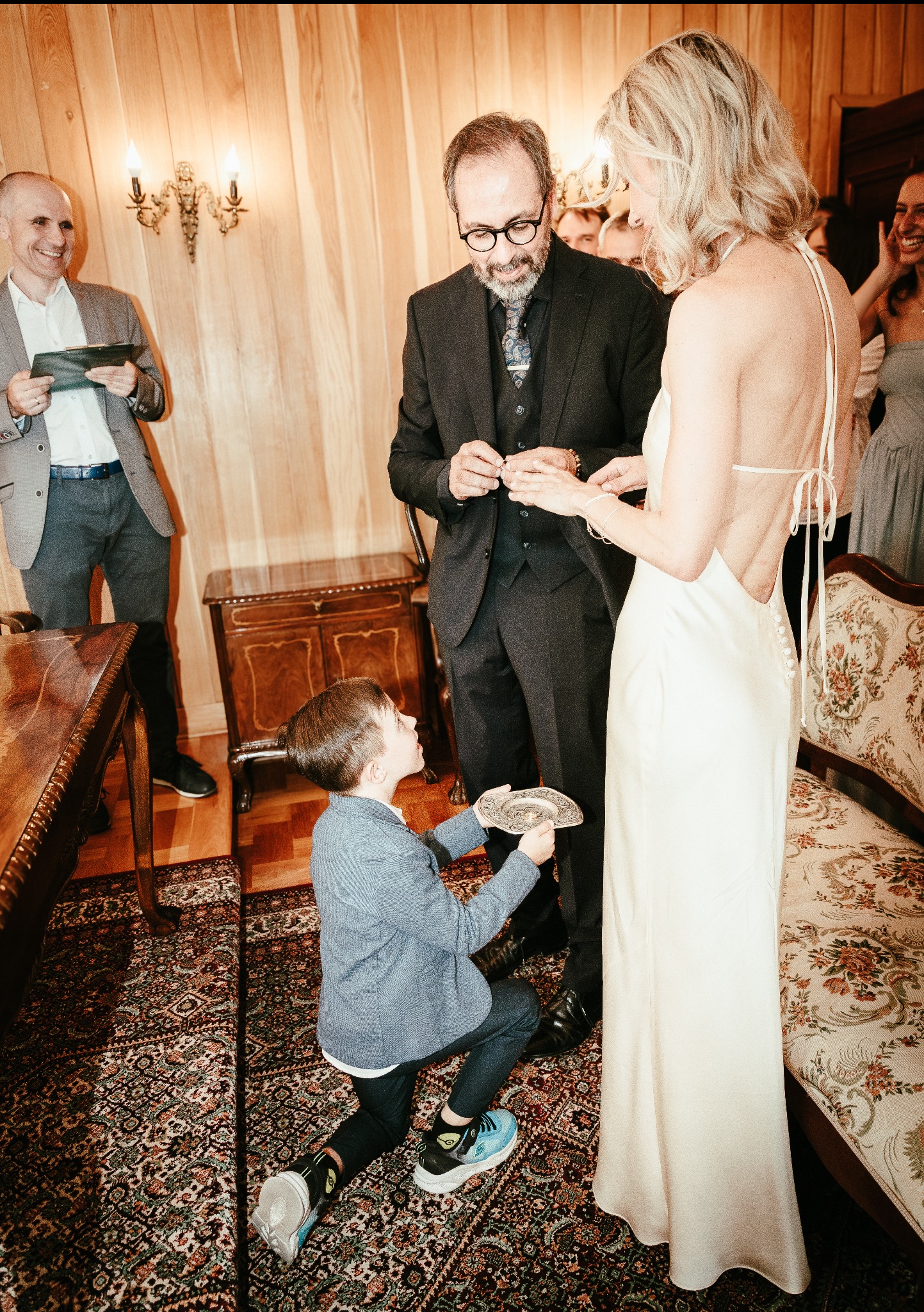 Chłopiec na kolanach podaje obrączki parze młodej podczas ceremonii ślubnej w pomieszczeniu z drewnianymi ścianami i wzorzystym dywanem. Goście w tle.