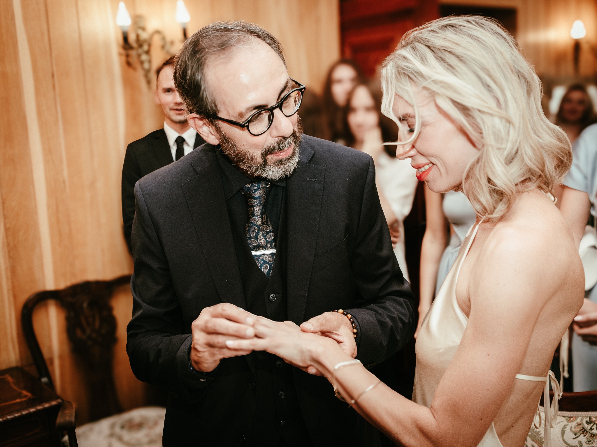 Elegancki mężczyzna w garniturze zakłada obrączkę uśmiechniętej kobiecie w białej sukience na tle gości weselnych w ciepłym oświetleniu sali.