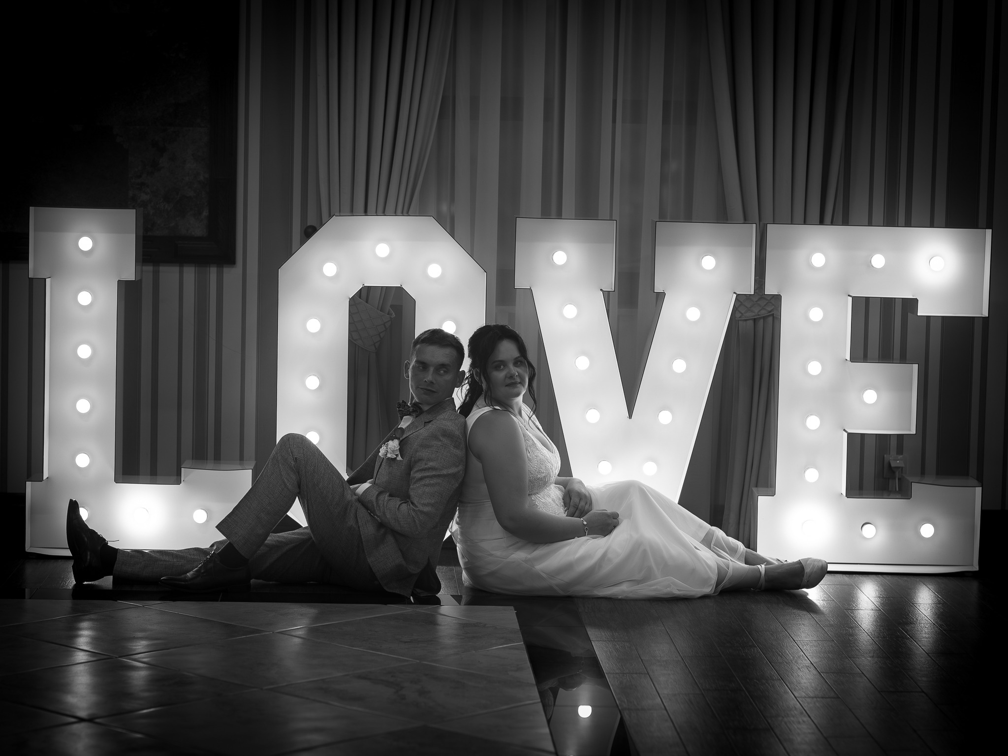 Czarno-białe zdjęcie pary młodej siedzącej plecami do siebie na podłodze, z napisem LOVE podświetlonym żarówkami w tle. Elegancki strój, subtelny uśmiech.