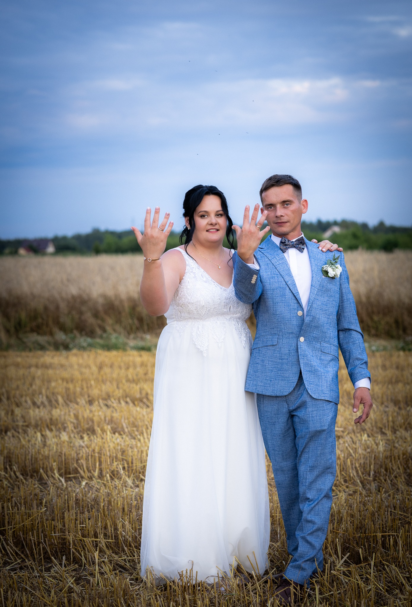 Para młoda prezentuje obrączki na tle pola po żniwach. Pan młody w błękitnym garniturze, panna młoda w białej sukni. Romantyczna sesja w plenerze.