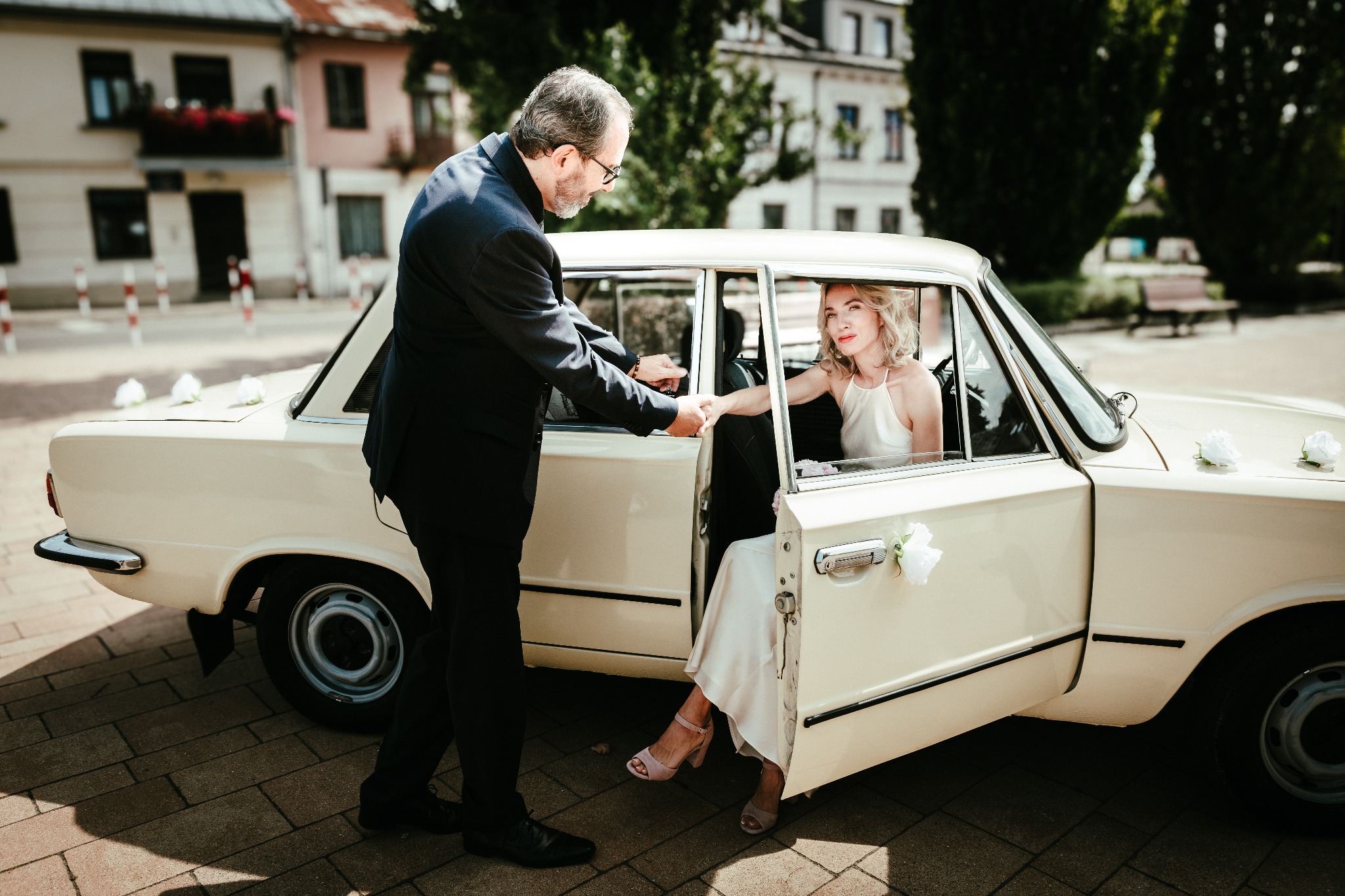 Panna młoda w beżowej sukni wysiada z kremowego, udekorowanego białymi różami samochodu, pomagający jej mężczyzna w garniturze. W tle budynki.