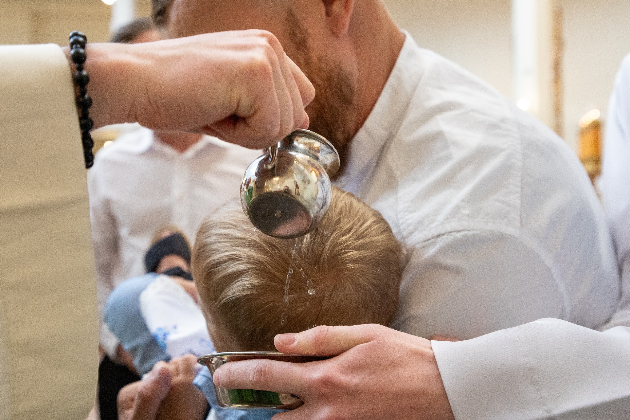 Ksiądz polewa wodą głowę dziecka podczas chrztu, widoczny dzbanek i miseczka na wodę, w tle osoby w białych koszulach.