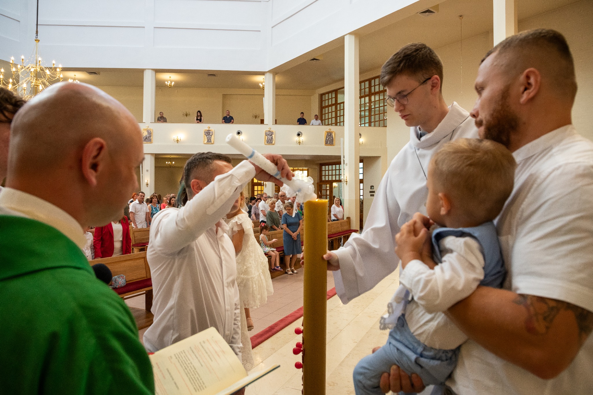 Uroczystość w kościele: ojciec chrzestny zapala świecę trzymaną przez księdza, dziecko w niebieskiej kamizelce obserwuje z rąk drugiego mężczyzny, w tle zgromadzeni goście.