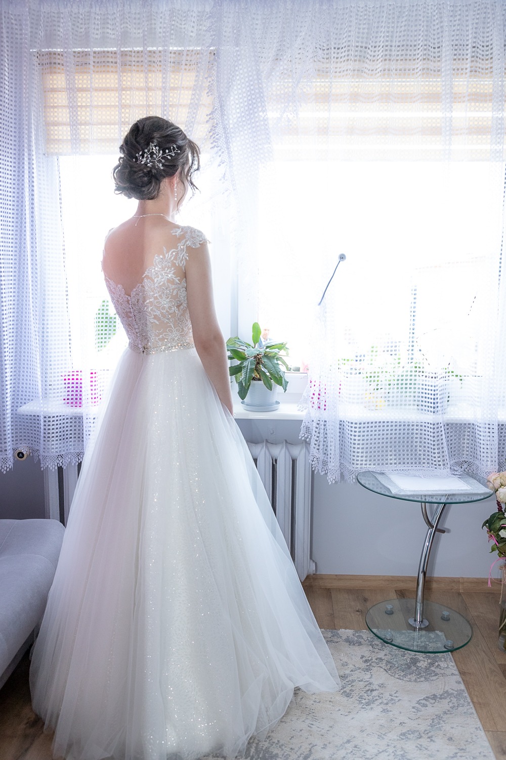 Panna młoda w białej sukni ślubnej z koronkowymi plecami, stojąca tyłem przy oknie z białymi firankami, z upiętymi włosami i ozdobą, delikatny makijaż, widok na jasne wnętrze pokoju.