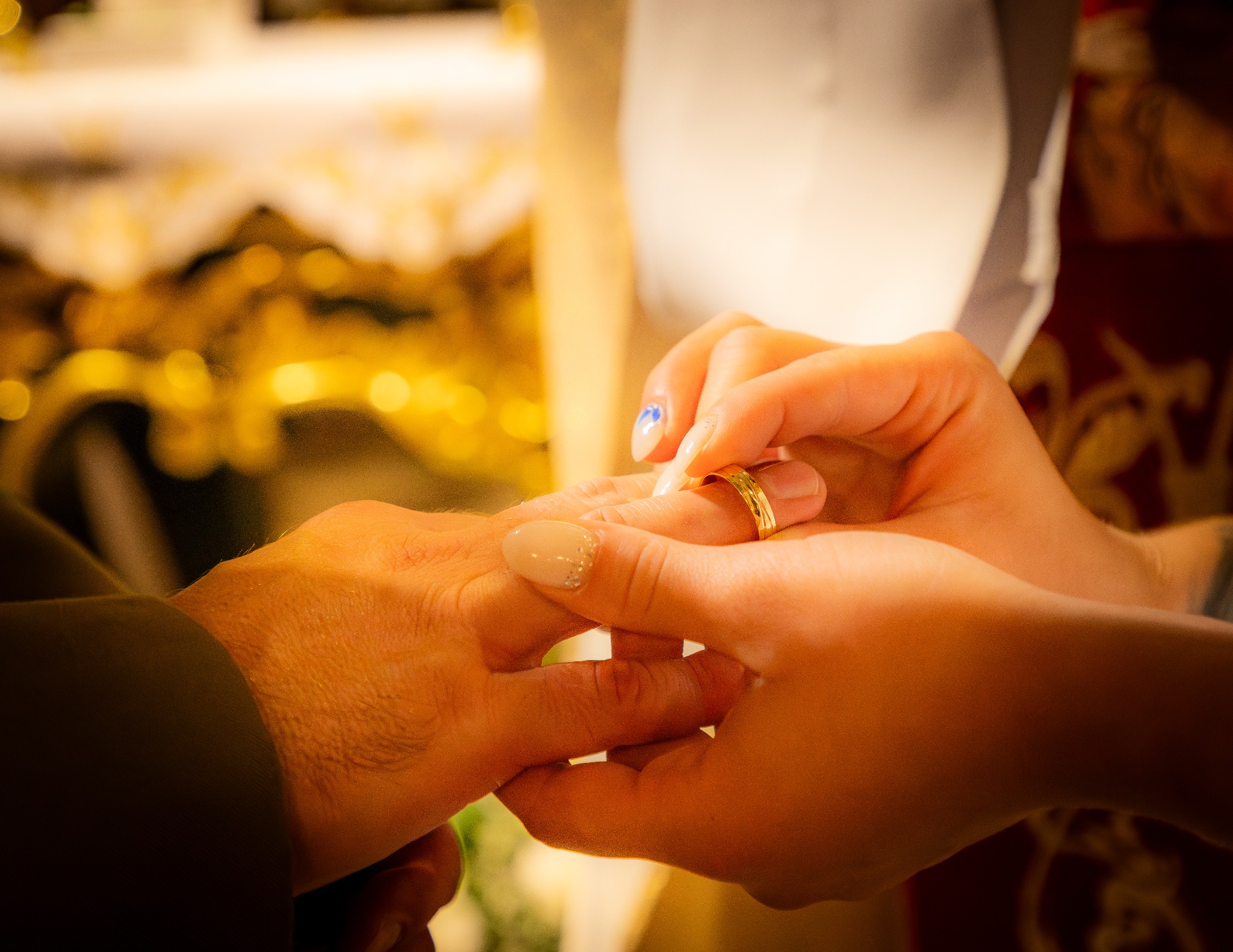 Zakładanie złotej obrączki na palec podczas ceremonii ślubnej, widoczne dłonie z manicure, ciepłe oświetlenie.