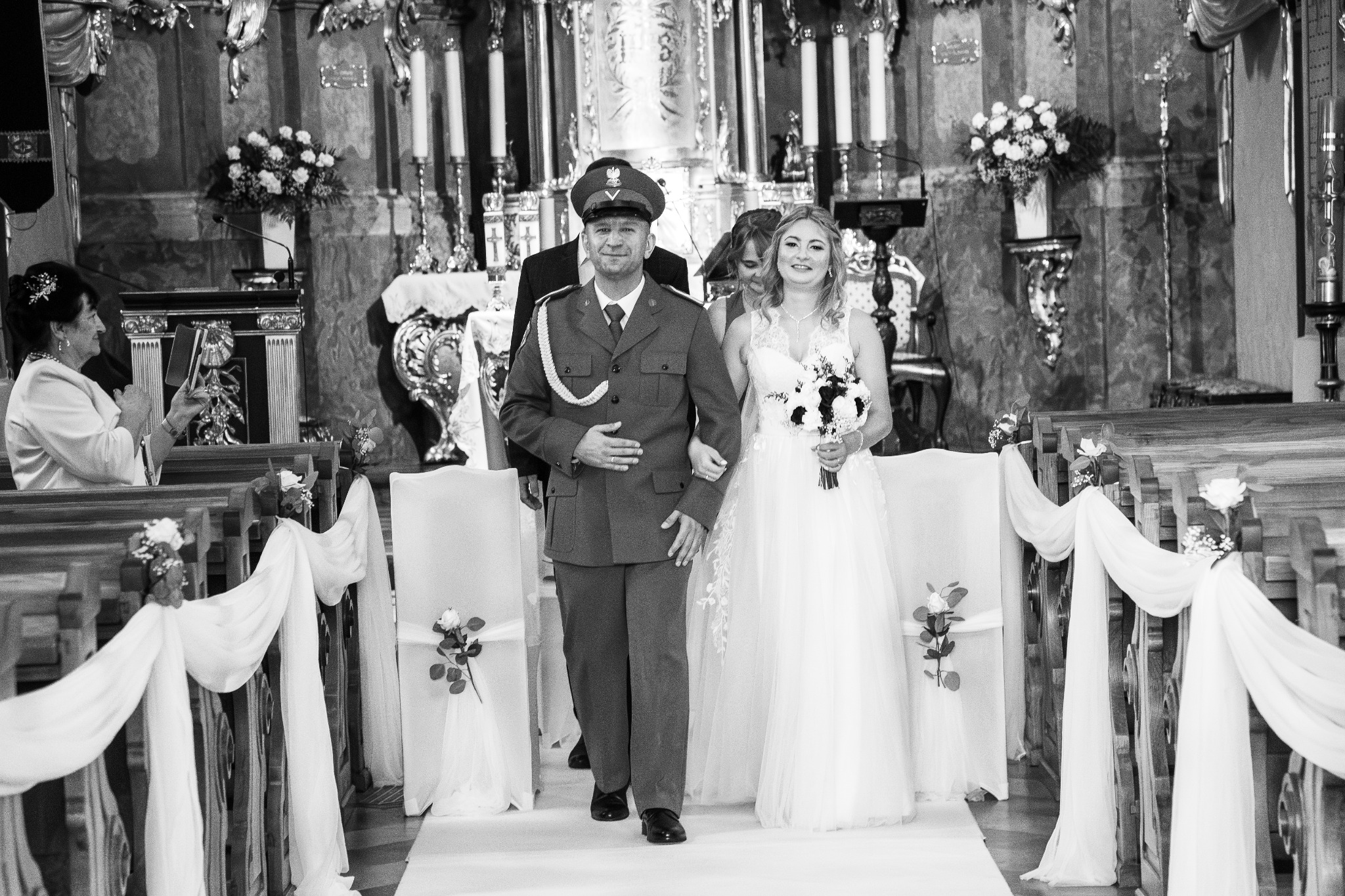 Czarno-białe zdjęcie panny młodej w białej sukni i mężczyzny w mundurze, idących w kościele udekorowanym białymi tkaninami i kwiatami.