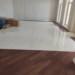 Proflazienka - Układanie podłogi: Połączenie białych płytek imitujących marmur z ciemnymi panelami drewnopodobnymi w przestronnym, jasnym wnętrzu z wysokimi oknami.