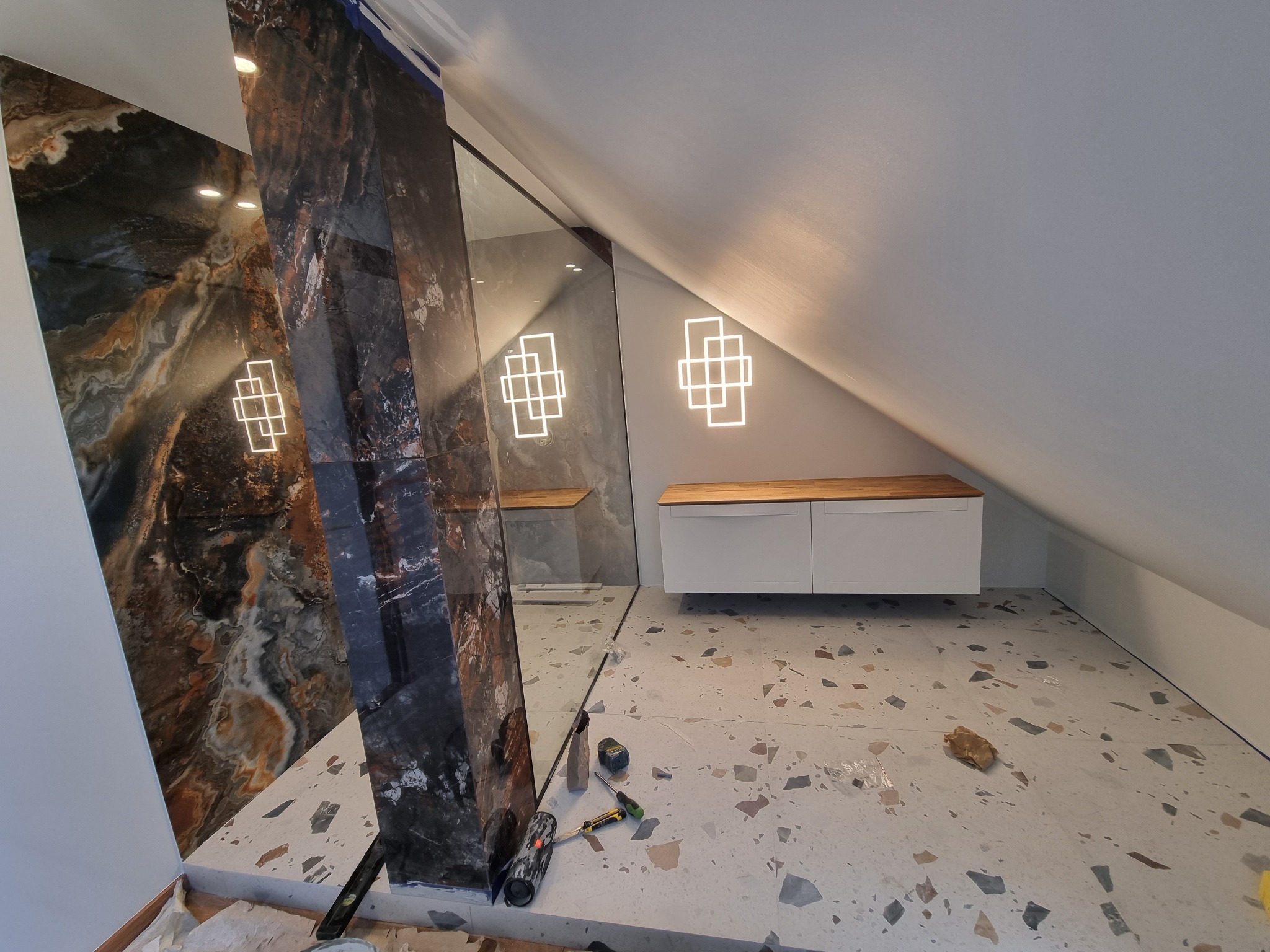 Montaż dużych, wzorzystych płytek w łazience na poddaszu w Katowicach, widoczne narzędzia glazurnicze, oświetlenie dekoracyjne i fragment szafki pod umywalkę.