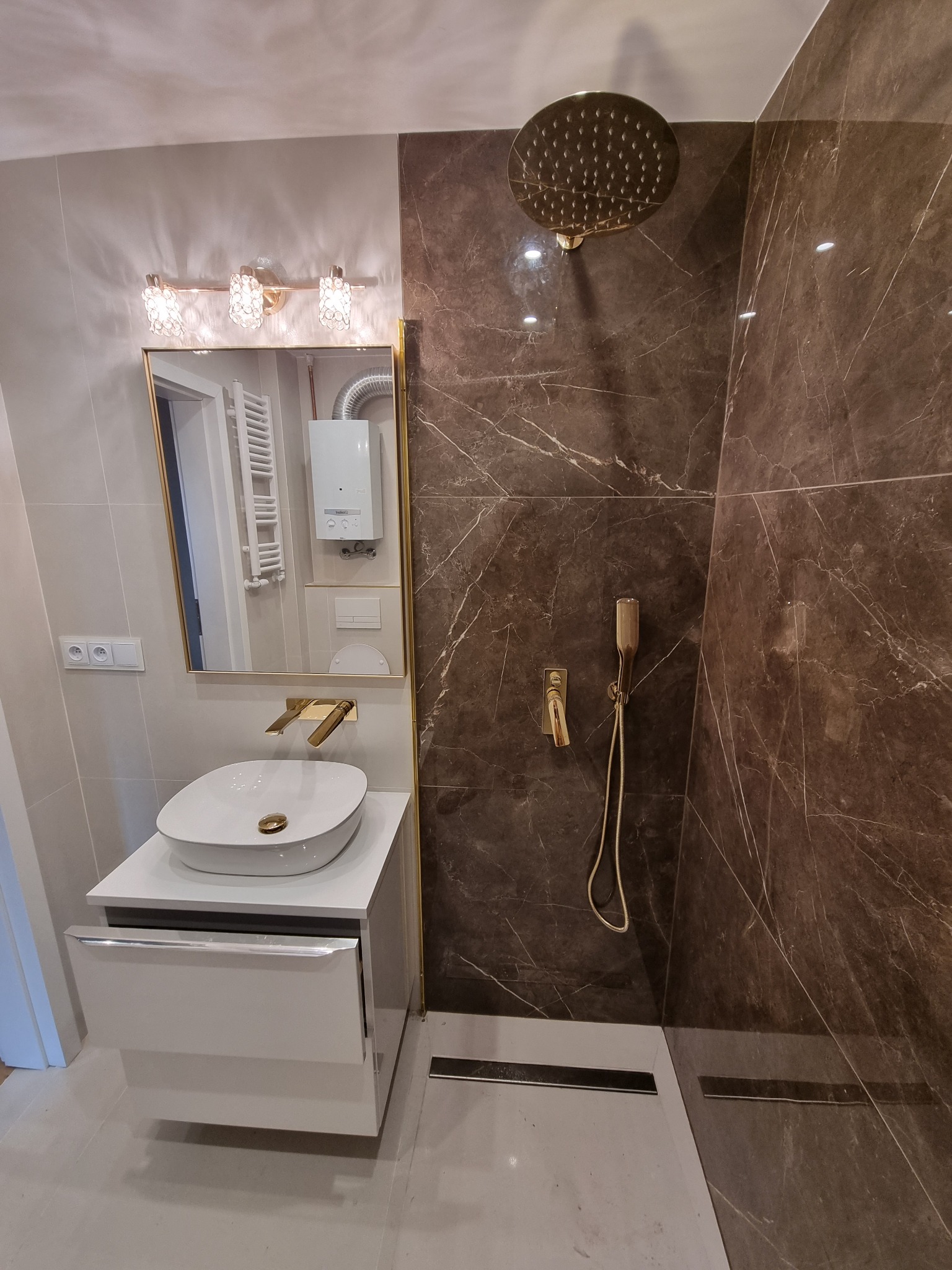 Nowoczesna łazienka z brązowymi płytkami imitującymi marmur, złotą armaturą i umywalką nablatową, oświetlona lampami kryształowymi