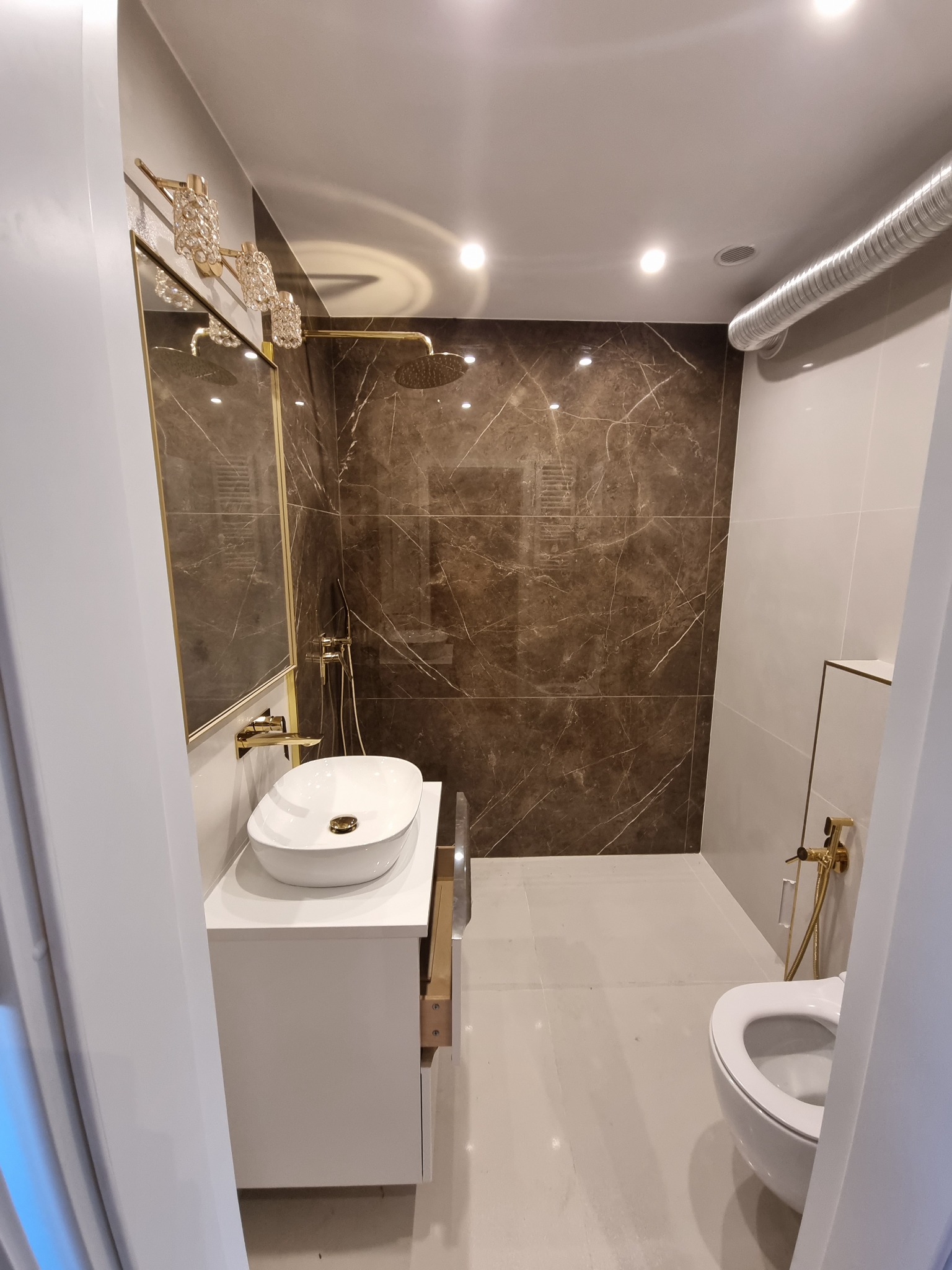 Elegancka łazienka z brązowymi płytkami imitującymi marmur, złotą armaturą, owalną umywalką nablatową i kryształowym oświetleniem.