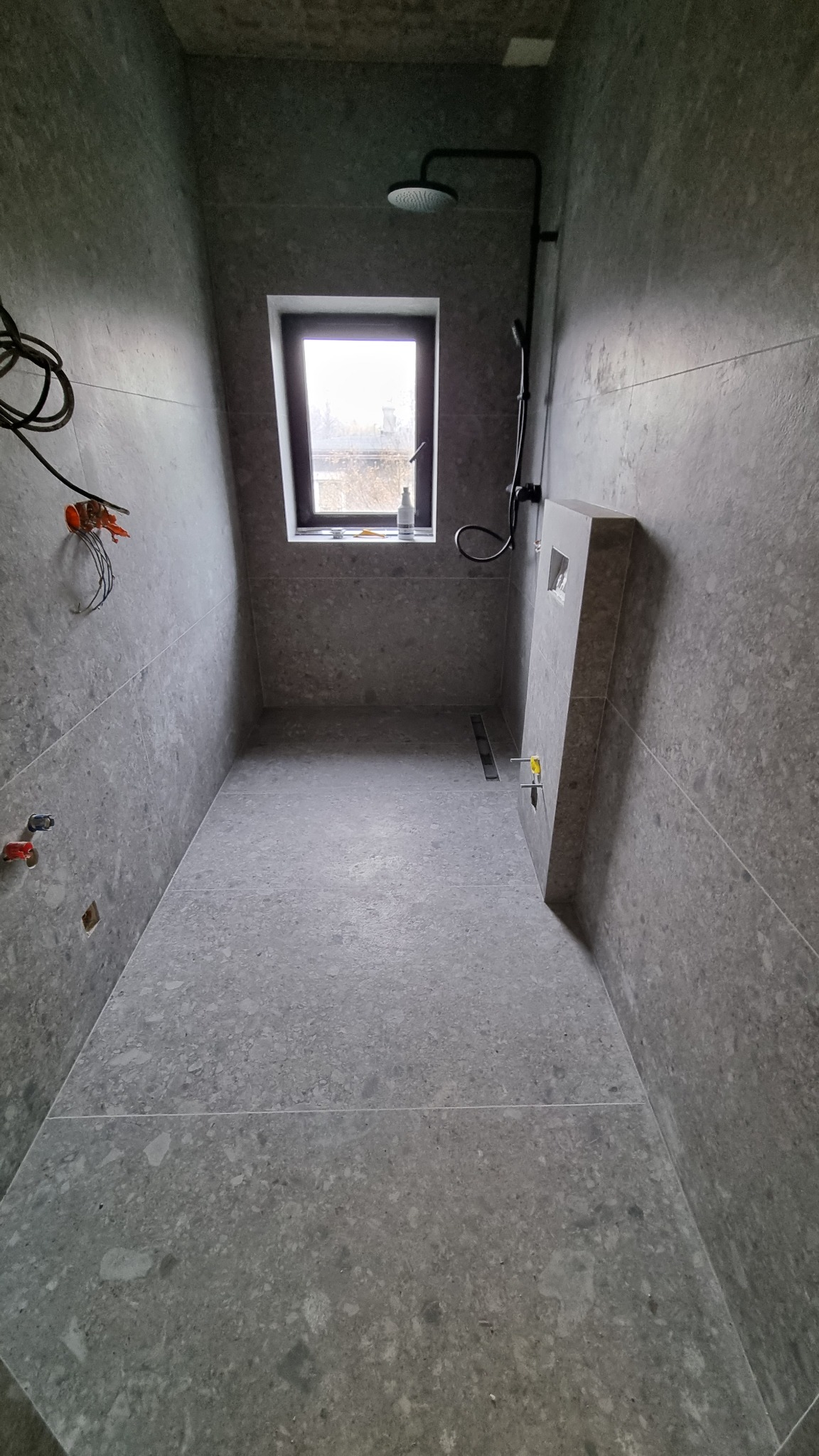 Wnętrze szarej łazienki z oknem, widoczne niedokończone instalacje elektryczne i hydrauliczne, nowoczesna deszczownica i odpływ liniowy.