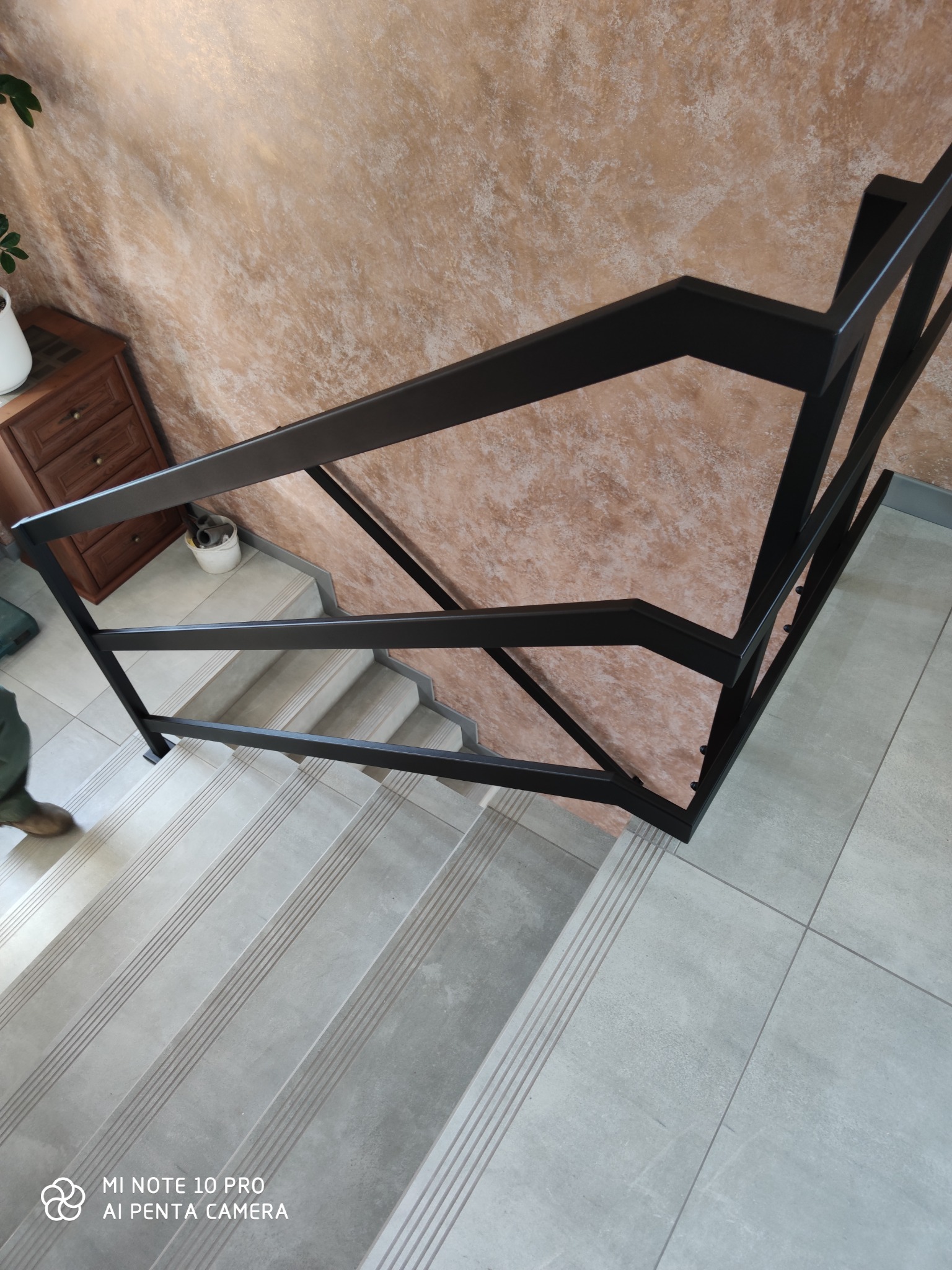 Kątowe ujęcie z góry na schody wyłożone szarymi płytkami z czarną, metalową balustradą o geometrycznym wzorze, z widoczną częścią buta osoby na schodach, na tle ściany o teksturze przypominającej...