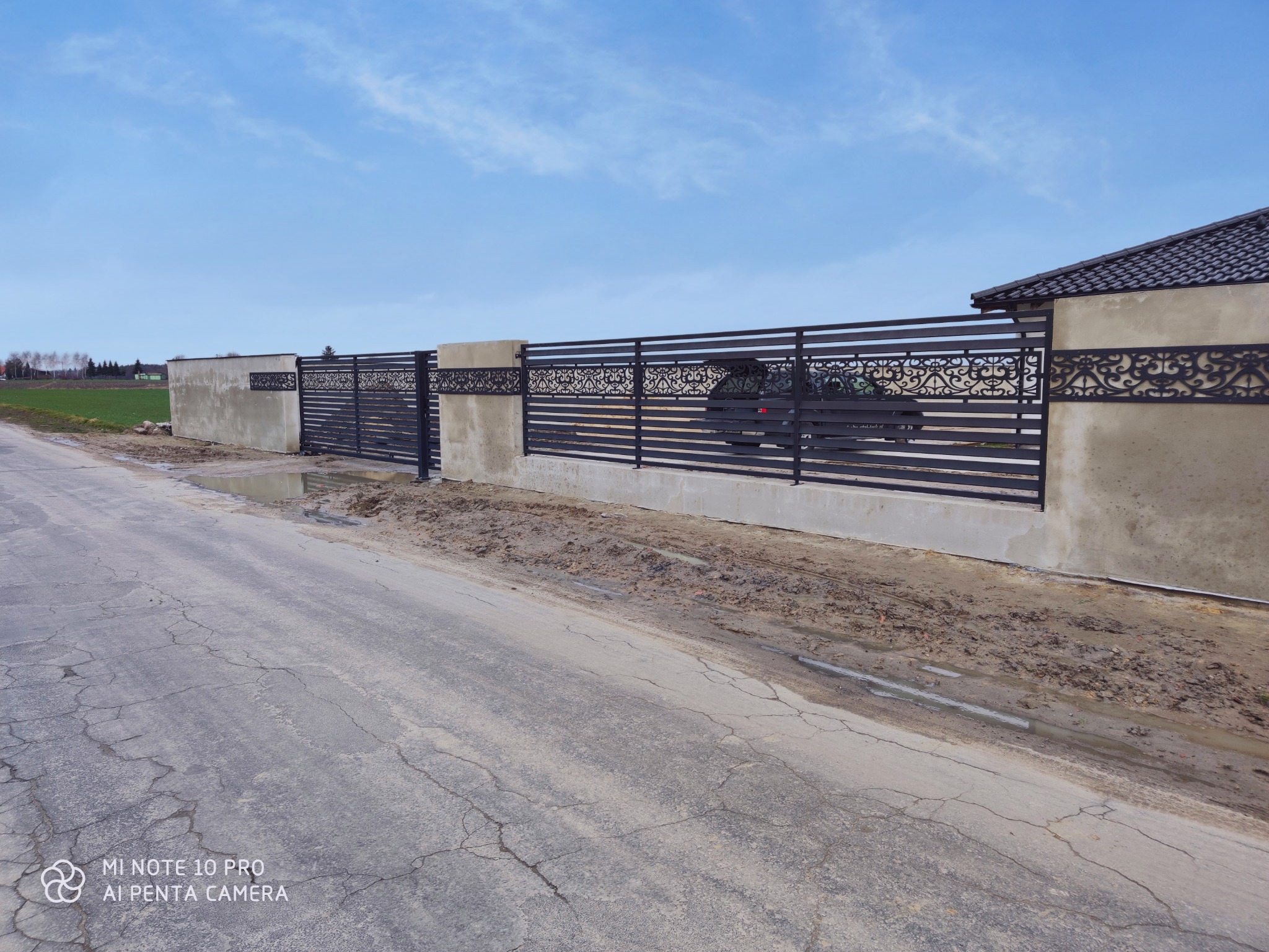 Nowoczesne ogrodzenie panelowe z elementami dekoracyjnymi i betonowymi słupkami, widok z ulicy, błoto po deszczu.