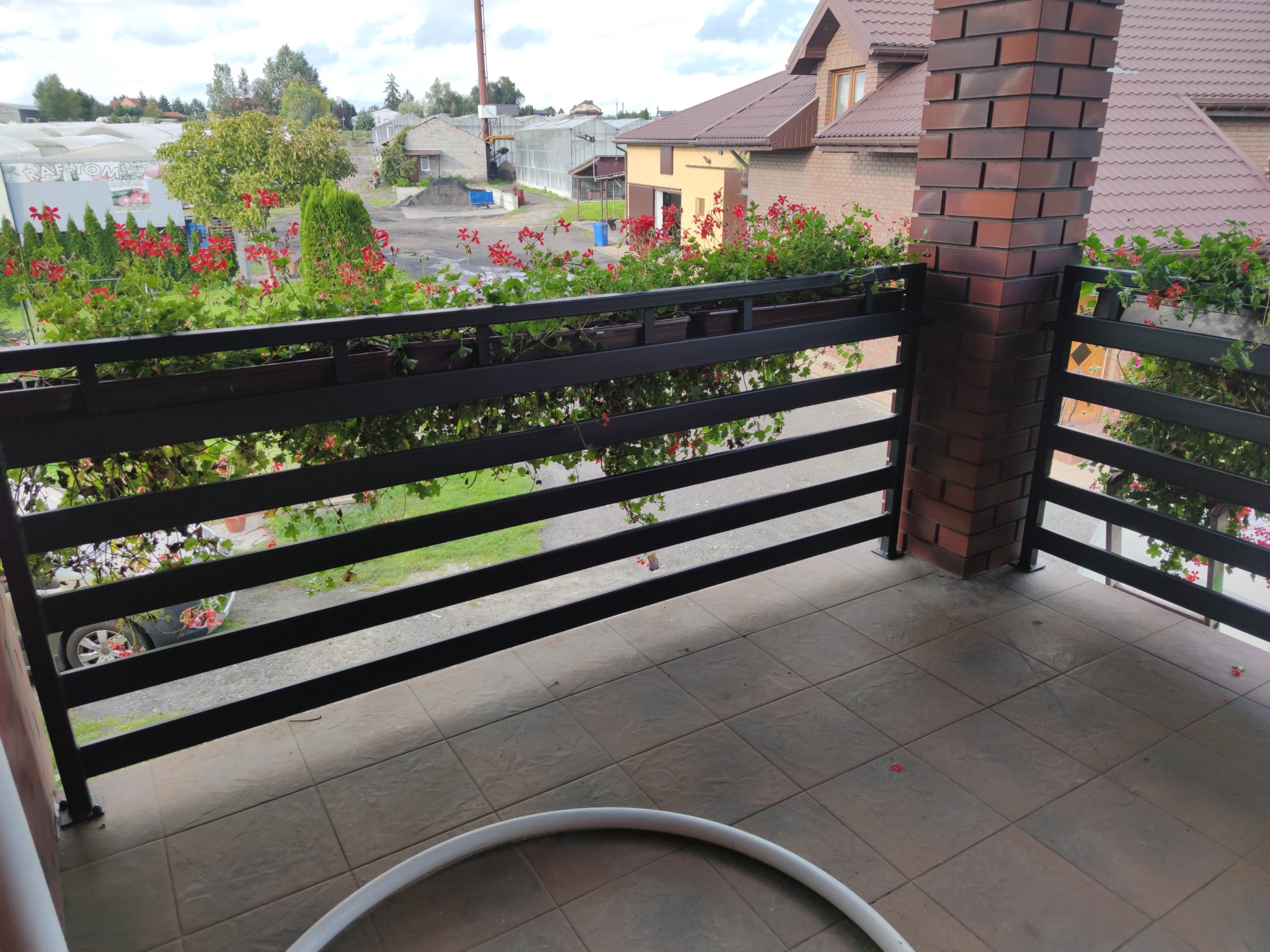 Czarna, pozioma balustrada balkonowa z kwiatami w donicach, wsparta na ceglanym filarze, widok z balkonu na budynki i zieleń w tle.