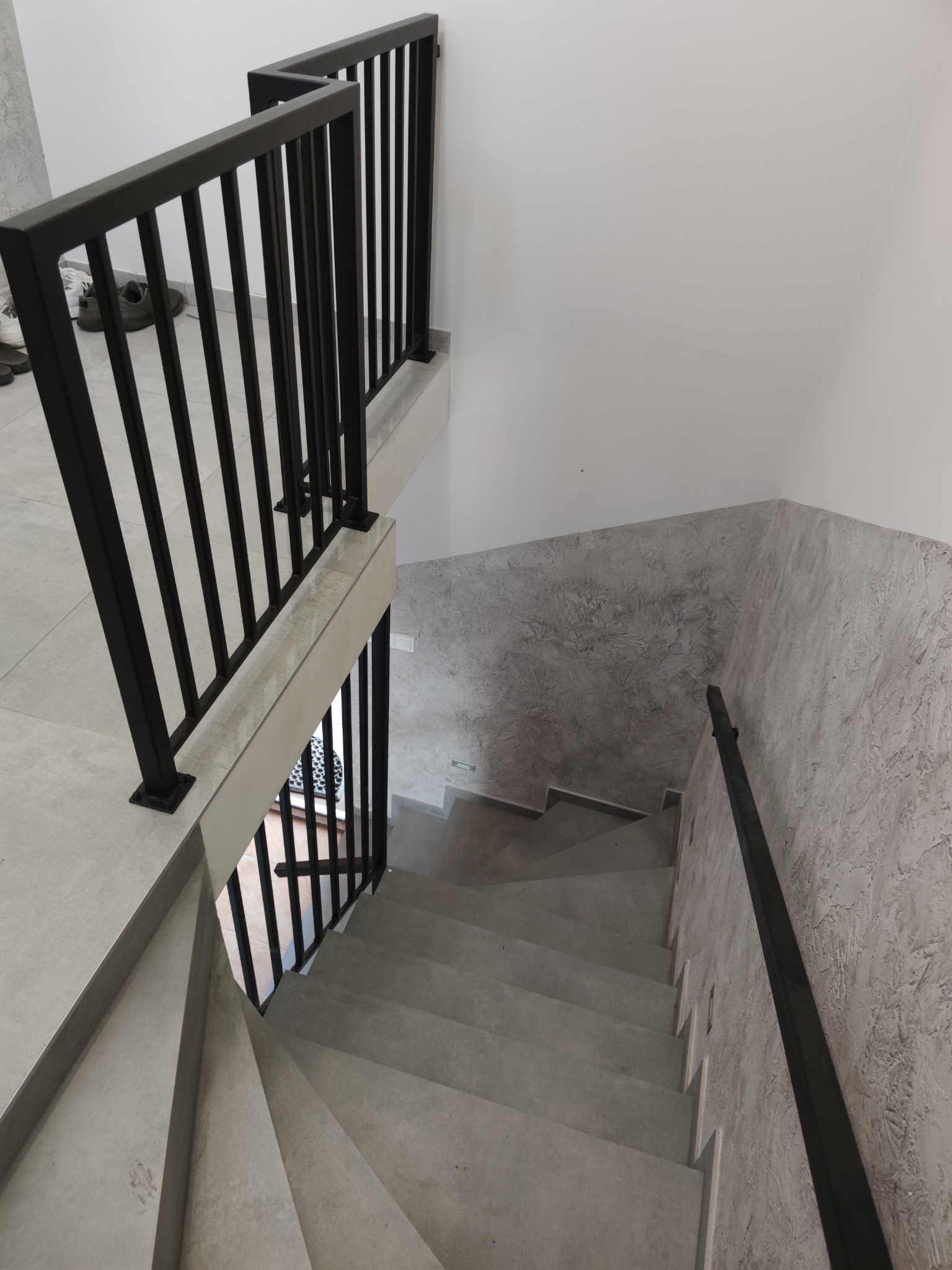 Wewnętrzne schody z szarymi stopniami i czarną, metalową balustradą, widok z góry, ściany wykończone dekoracyjnym tynkiem imitującym beton.