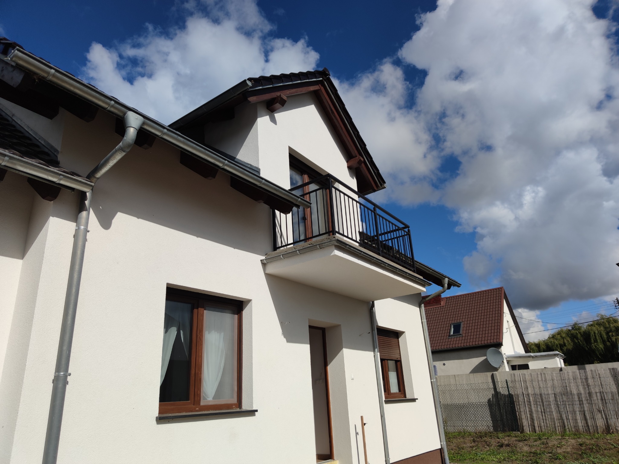 Nowoczesny dom jednorodzinny z balkonem z czarną metalową balustradą, widok z dołu na białą elewację i błękitne niebo z chmurami.