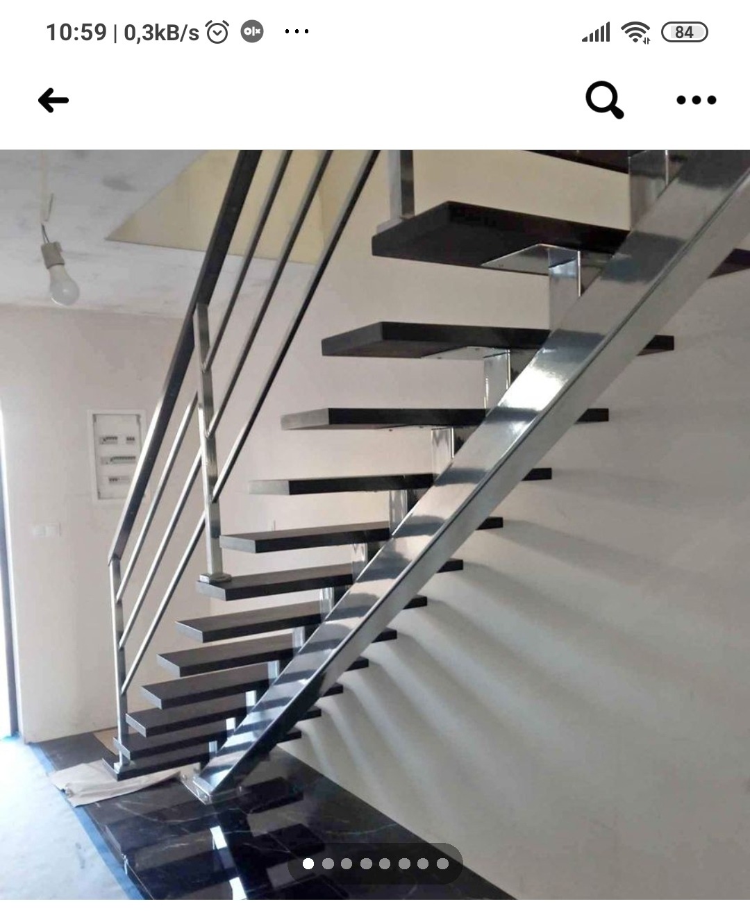 Wewnętrzne schody z czarnymi stopniami i metalową balustradą, widoczne z dołu, z marmurową podłogą i surowym oświetleniem.