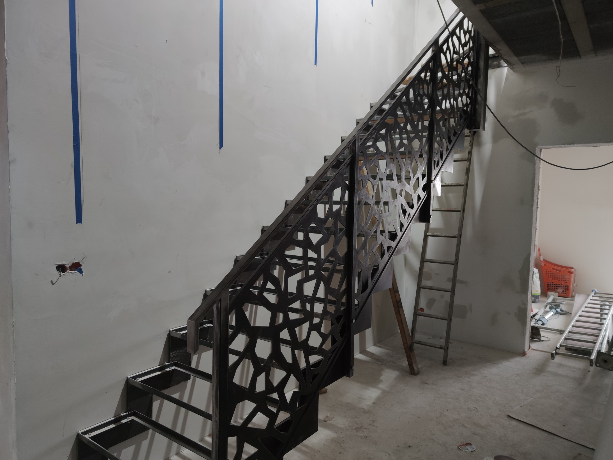 Konstrukcja schodów stalowych z ażurową balustradą o geometrycznym wzorze, w surowym stanie, w trakcie montażu wewnątrz budynku, widoczne ślady prac wykończeniowych, drabina oparta o ścianę.