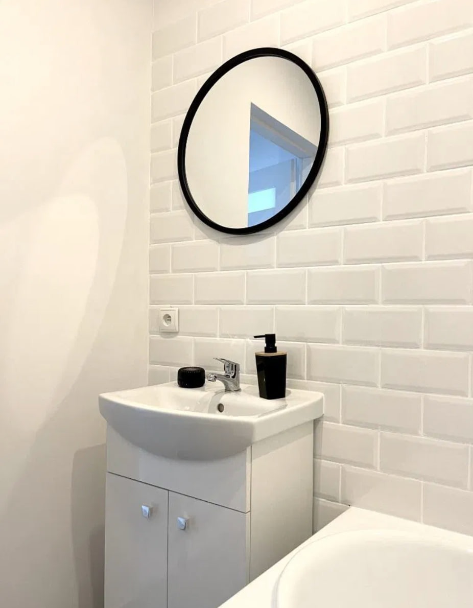 Biała łazienka z umywalką na szafce, okrągłym lustrem w czarnej ramie i białymi płytkami typu metro na ścianie, z widocznym fragmentem wanny i dozownikiem mydła.