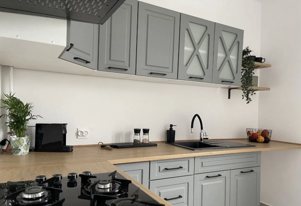 Nowoczesna kuchnia w odcieniach szarości z drewnianym blatem, czarną armaturą i płytą gazową. Górne szafki z przeszkleniami w kształcie litery X. Na półce roślina doniczkowa.