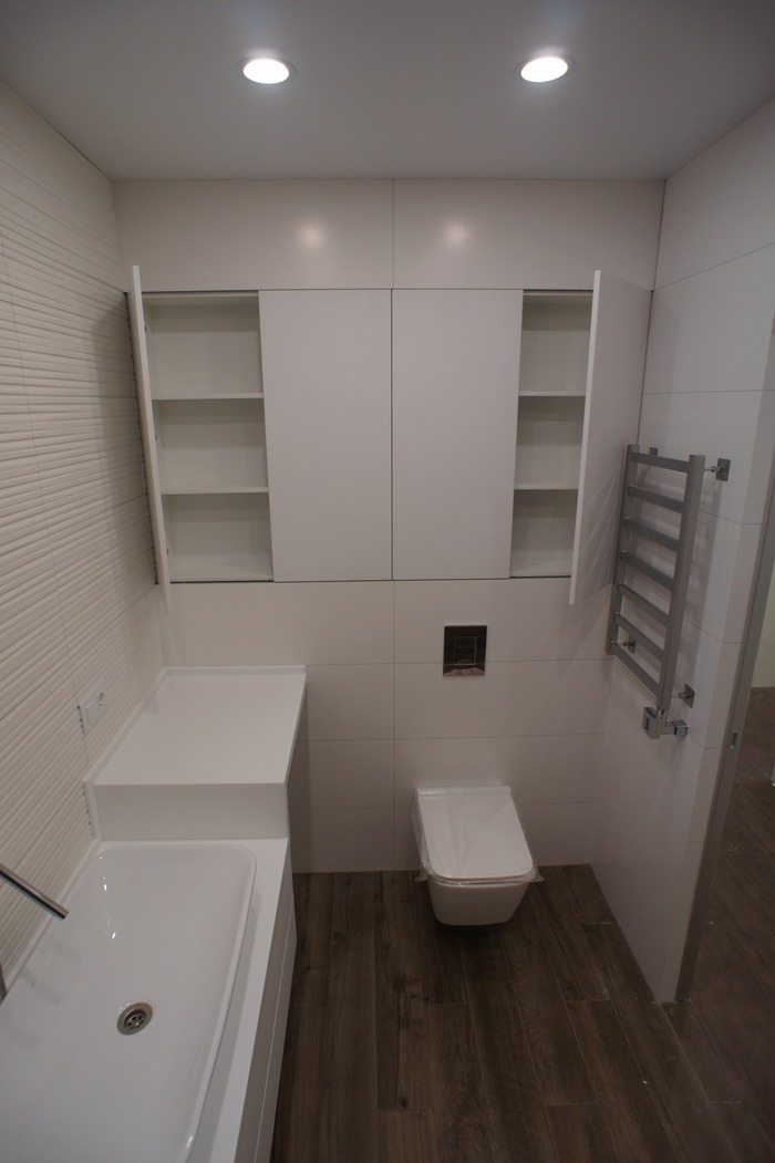 Nowoczesna biała łazienka z zabudową meblową nad toaletą i umywalką, grzejnik drabinkowy na ścianie, podłoga imitująca drewno, jedna szafka otwarta.