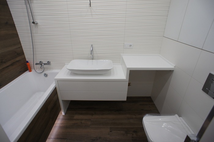Nowoczesna łazienka z białymi płytkami o falistej strukturze, drewnopodobną obudową wanny, białą umywalką nablatową na szafce i drewnopodobną podłogą.