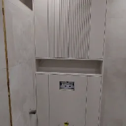 Szara zabudowa meblowa w łazience z szafkami górnymi z pionowymi żłobieniami, półką i dolną szafką z widocznym otworem na stelaż podtynkowy WC.