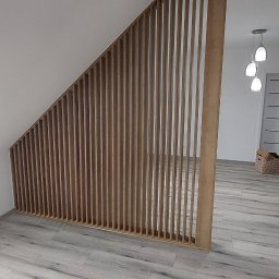 MEL MEBLE Mateusz Leszczyński - Drewniana konstrukcja z pionowych listew, tworząca ażurową ściankę działową w jasnym wnętrzu z podłogą imitującą drewno, widok na dalszą część pomieszczenia z lampami wiszącymi.