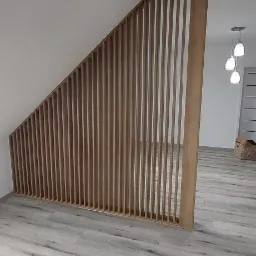 Drewniana konstrukcja z pionowych listew, tworząca ażurową ściankę działową w jasnym wnętrzu z podłogą imitującą drewno, widok na dalszą część pomieszczenia z lampami wiszącymi.