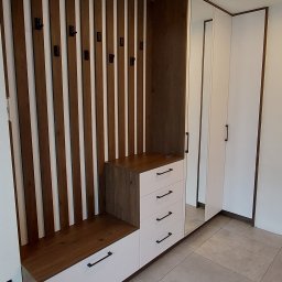 MEL MEBLE Mateusz Leszczyński - Wnęka z siedziskiem i wieszakami z pionowych listew drewnianych, połączona z białą szafą z lustrem, czarne uchwyty szuflad i drzwi.