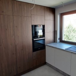 MEL MEBLE Mateusz Leszczyński - Nowoczesna kuchnia z frontami w odcieniach brązu, zabudowana piekarnikiem i mikrofalówką marki Bosch, jasny blat z oknem w tle, widoczna zieleń drzewa za oknem.