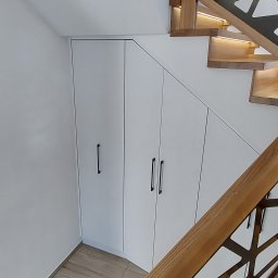 MEL MEBLE Mateusz Leszczyński - Biała zabudowa pod schodami z trzema frontami i minimalistycznymi, czarnymi uchwytami, dopasowana do kąta nachylenia schodów z drewnianymi stopniami z podświetleniem LED.