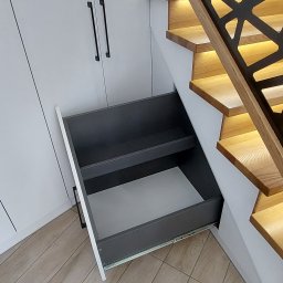 MEL MEBLE Mateusz Leszczyński - Wysuwana szuflada z półkami, wbudowana pod drewnianymi schodami z podświetleniem LED, obok białe szafki z czarnymi uchwytami.