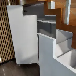 Biała szafa wnękowa pod schodami z otwartymi drzwiczkami, widoczne półki wewnątrz, obok drewniane schody z metalowymi elementami.